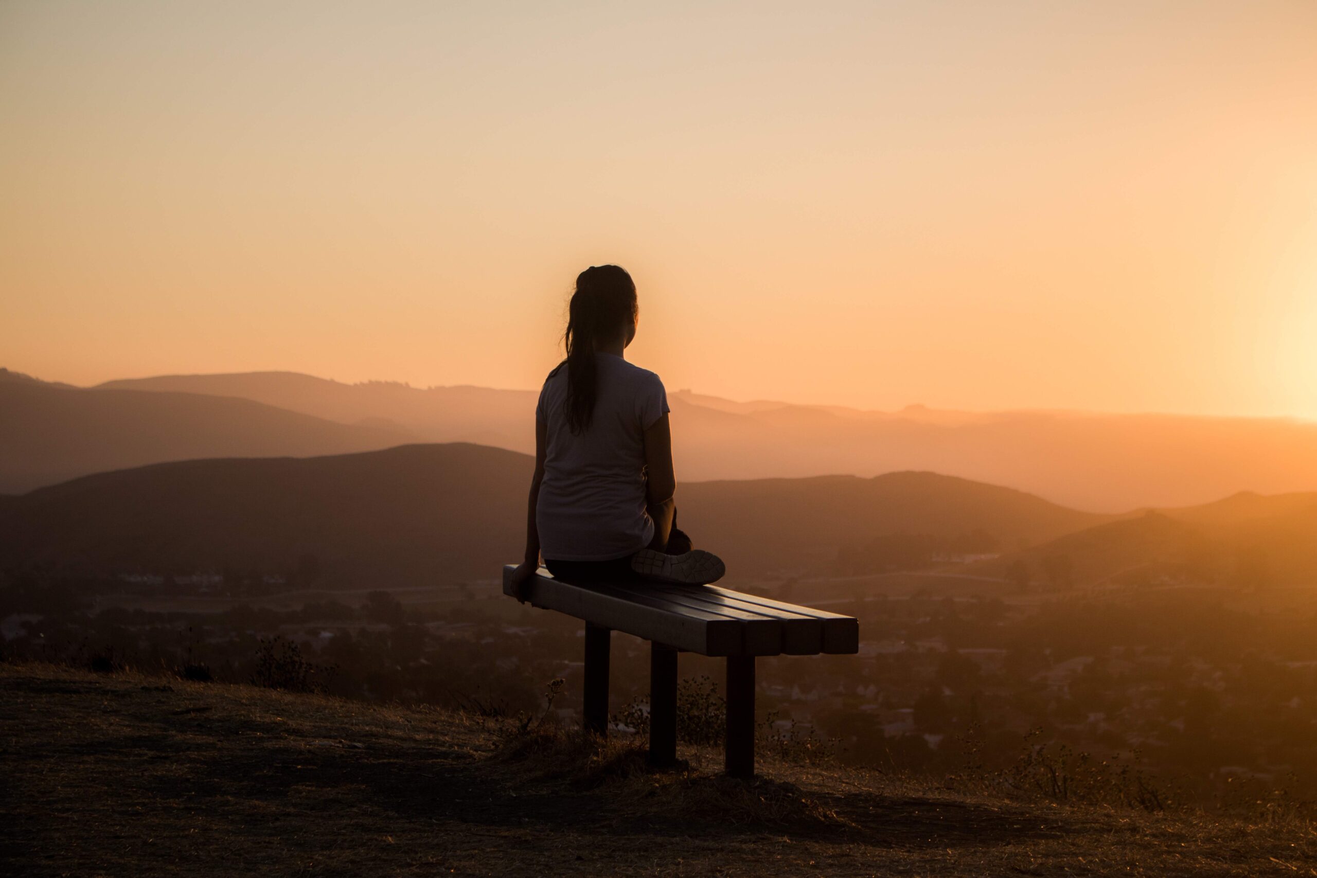 a girl sitting alone on a wooden bench enjoying sunset view.